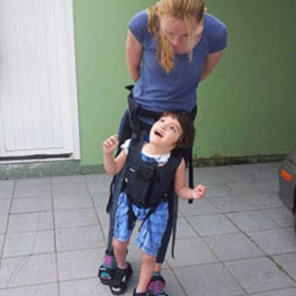 A child smiles up at an adult. The child is being supported by the Upsee harness, attached to the adult.