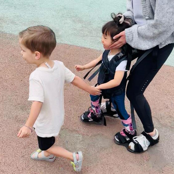 Mother and two children outdoors. The boy is running whilst holding the girl's hand. The girl is being supported by the Upsee harness, attached to her mother