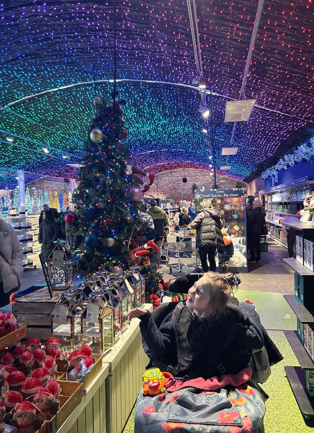 A little girl in a wheelchaiar smiles in a shop surrounded by Christmas decorations