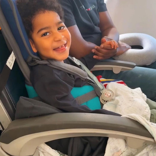 Niño sonriendo a la cámara, mientras está apoyado en su asiento GoTo a bordo de un avión. Su padre, fuera de plano, le coge de la mano.