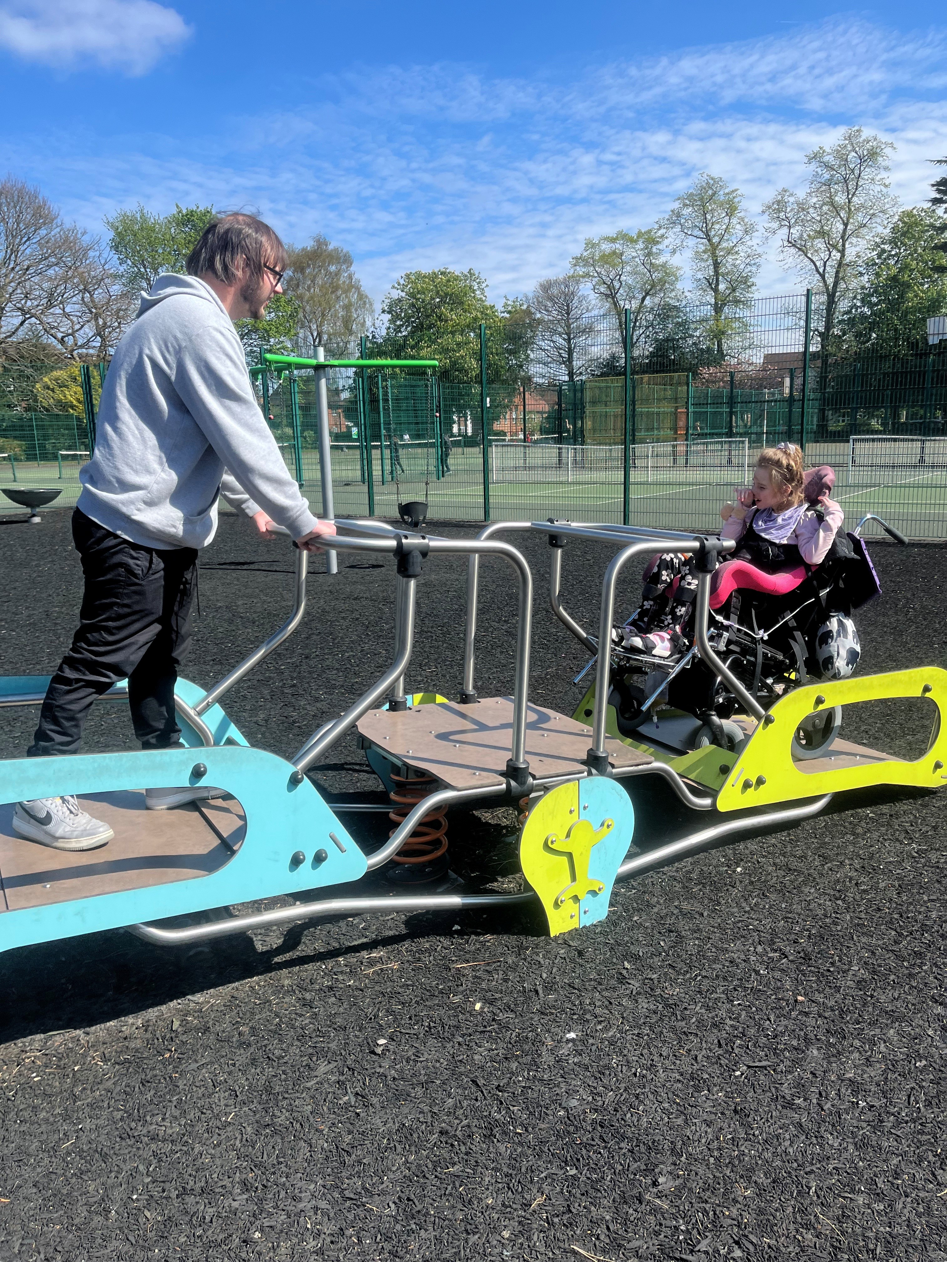A young girl in a wheelchair uses an accessible seesaw with her father.