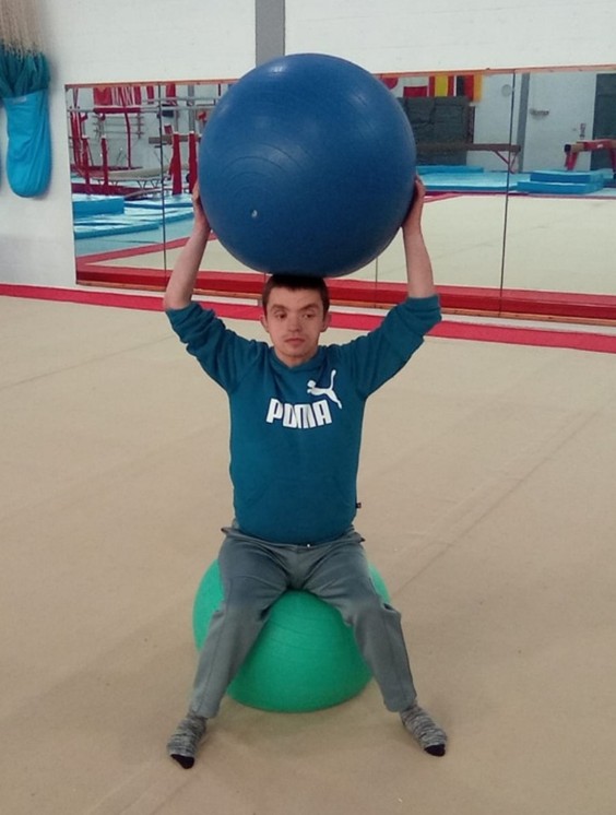 A young man sitting on a yoga ball, holding another larger yoga ball over his head.