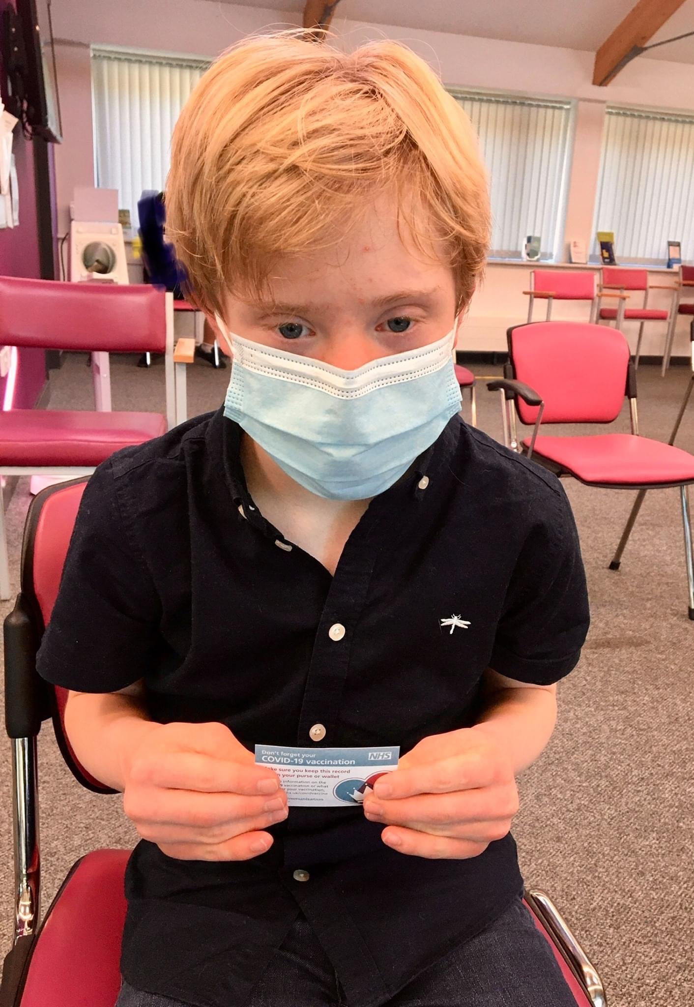 A young boy sits with a face covering on holding a vaccination card.