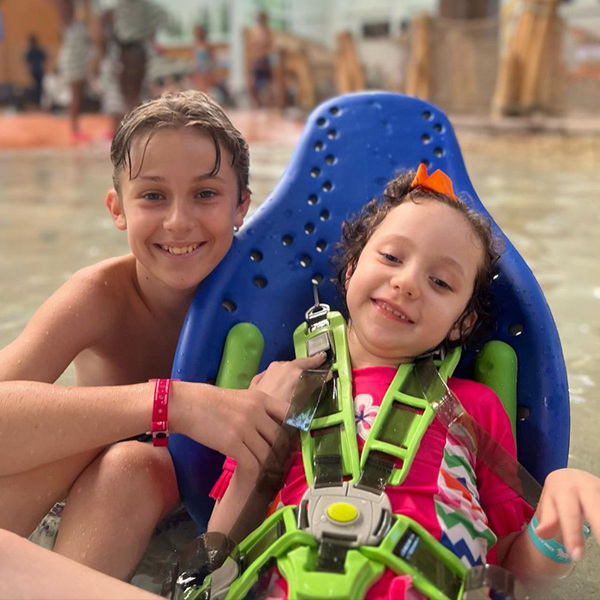 Siblings at a waterpark. One sits supported in her Splashy Big multipurpose bath seat.