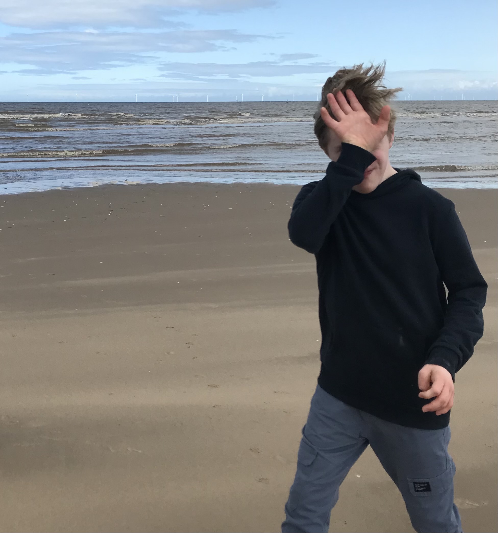 A young man walks along the beach while covering his face with the back of his hand.