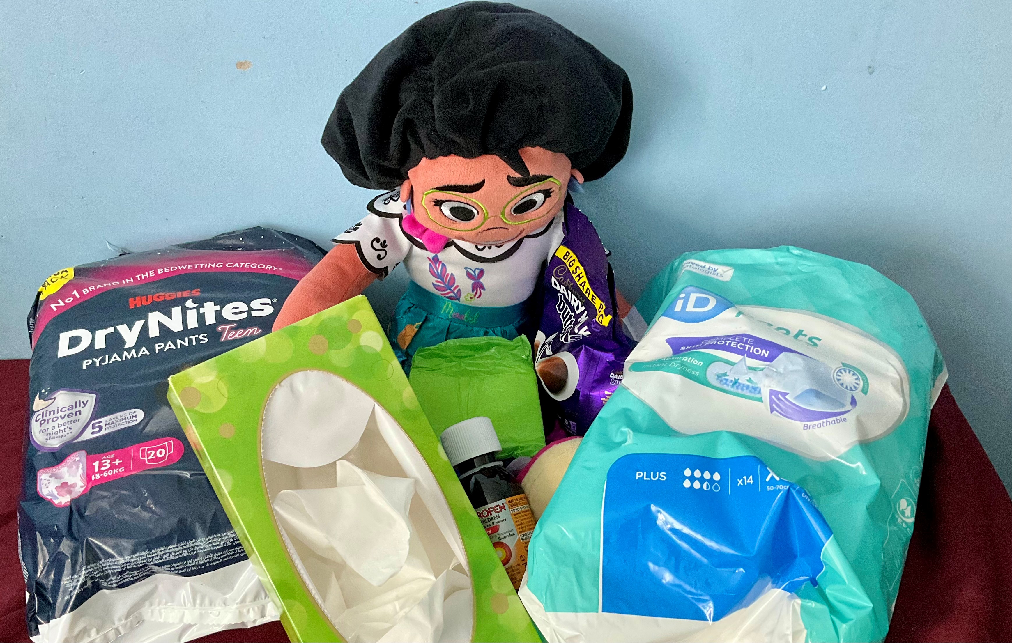 A Disney plush doll surrounded by period and incontinence products along with pain killers, tissues and chocolate