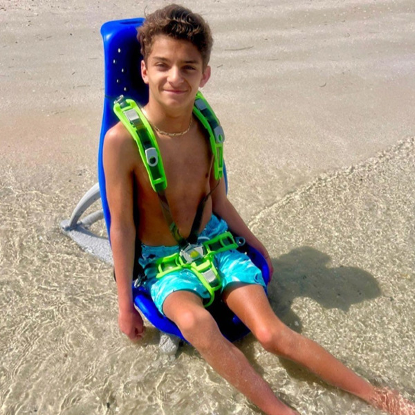 A boy on the beach sits in a Splashy Big multipurpose bath seat.