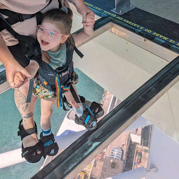 Child standing on a glass floor, supported by the Upsee mobility harness and being held by their parent