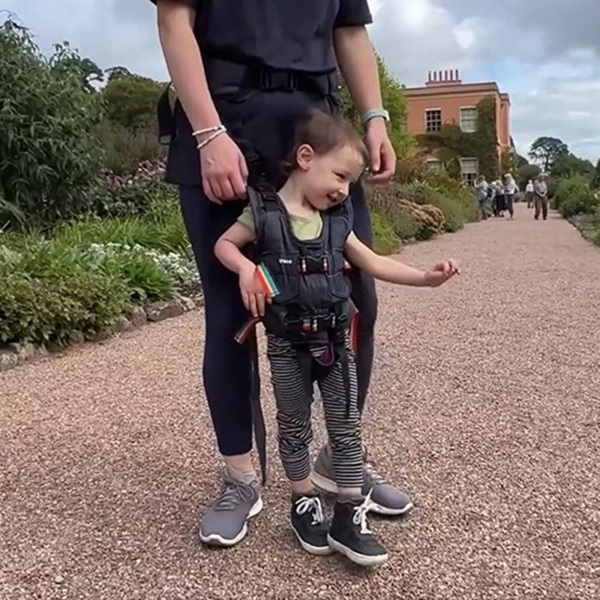 Niño al aire libre, apoyado en el arnés de movilidad Upsee en un paseo por jardines.