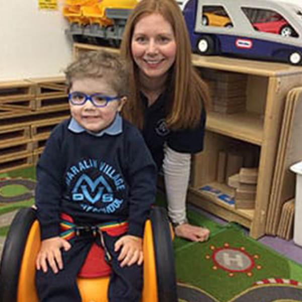 A little boy smiles in his Scooot sit and ride while his teacher sits beside him.