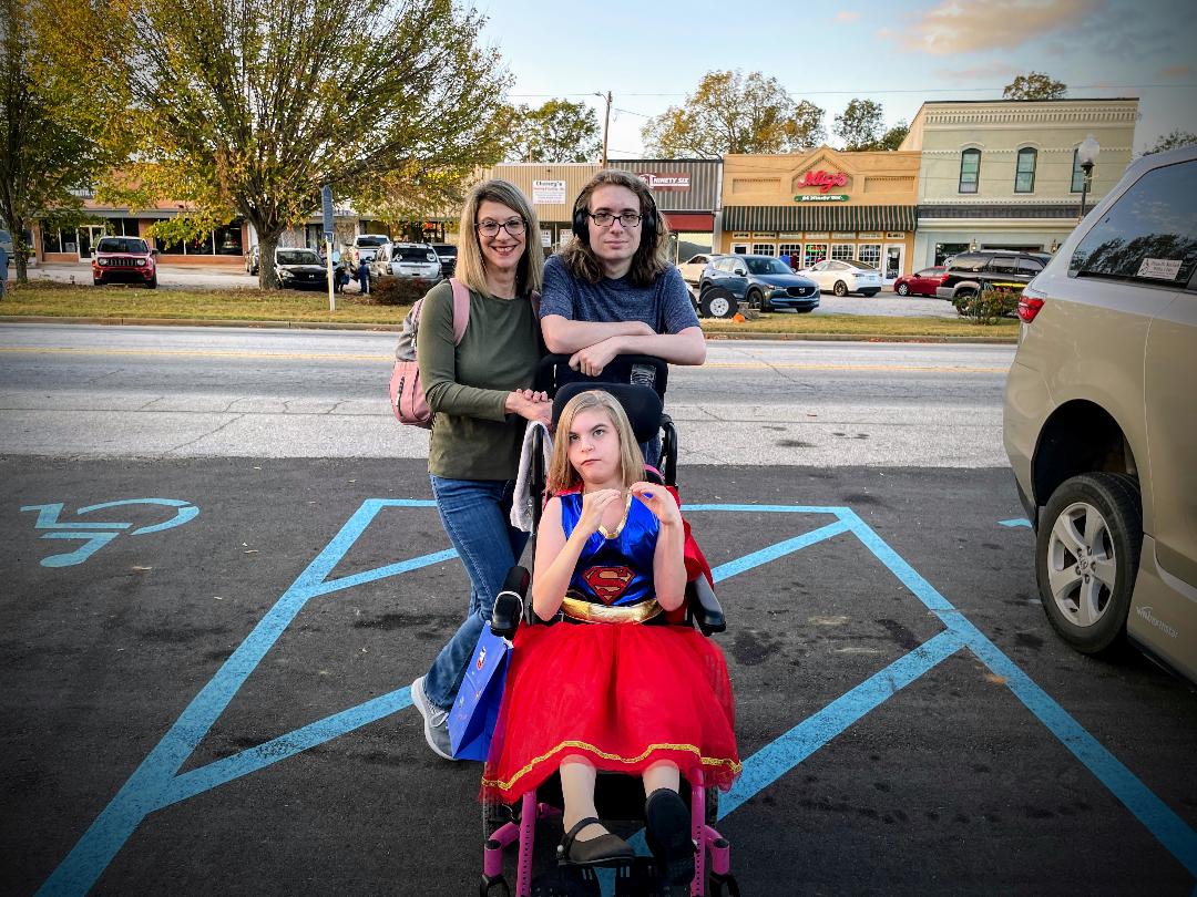 A family photo of mother, son and daughter. The daughter is dressed in a Superwoman costume in her wheelchair.